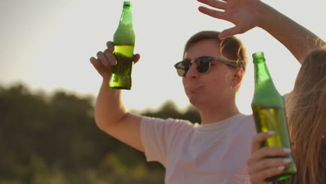 A-brutal-young-man-is-dancing-in-trendy-black-sun-glasses-and-white-T-shirt-with-beer-on-the-open-air-party-with-a-beautiful-young-blonde-girl-with-long-hair-in-red-plaid-shirt.